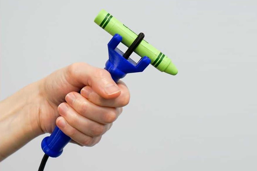 Functionalhand Universal Cuff device in blue as held by a person's left hand with a green crayon attached on the tip of the device and fasten with black elastic cord.