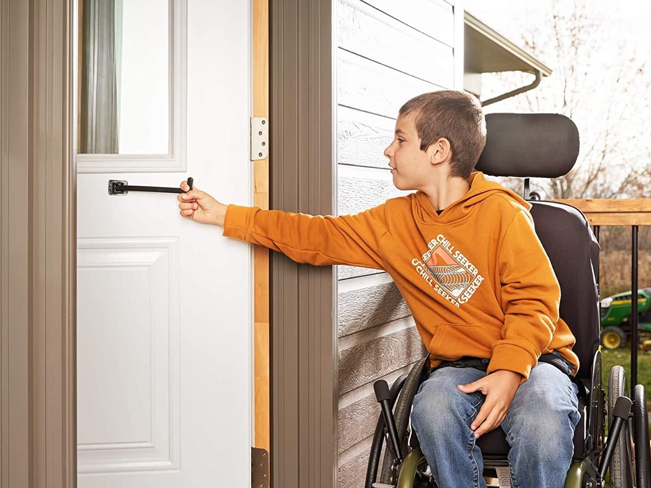Shows a boy on wheelchair pulling the handle of the T-Pull Door Closer device which is fixed on a door at a level which enables him to close the door easily after he goes through it with his wheelchair.