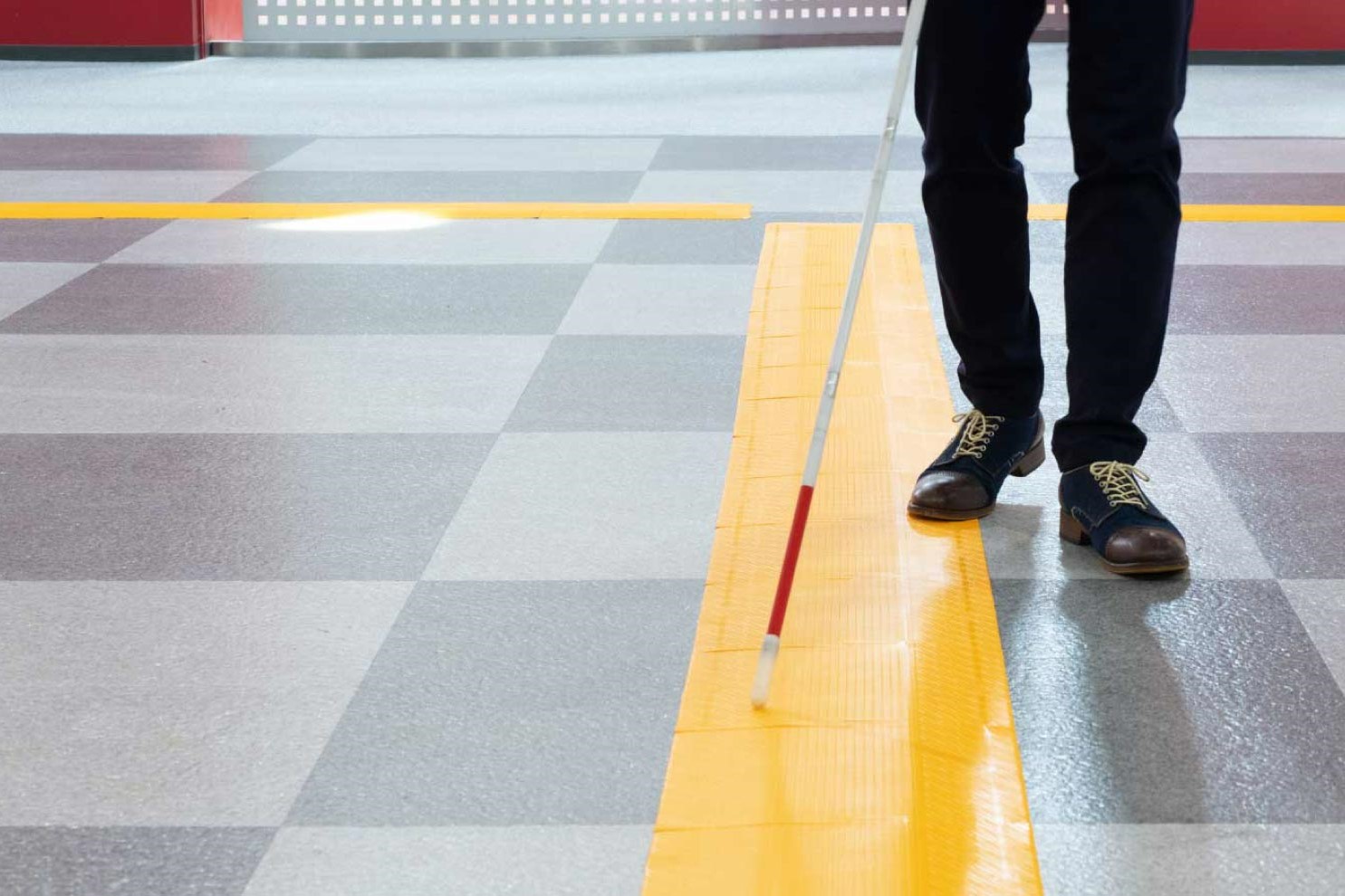 A stretch of the Hodokun guideway rubber mats in yellow shown on the floor. A man is shown stepping on the edge of the mats and using his white cane to feel around while he navigates his way.