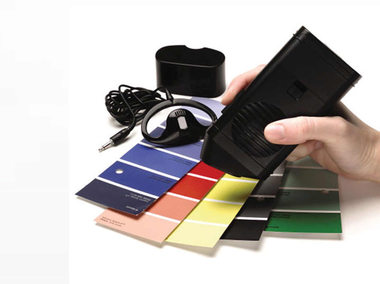 Talking Colour Detector held by a person's hand, with five cards of colour samples in the shades of blue, red, yellow, black and green placed in front of the device.
